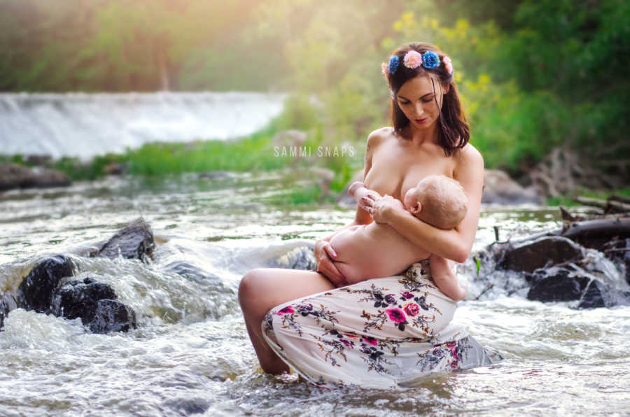 Mother sits on rock in river with baby in her lap who is breasfeeding