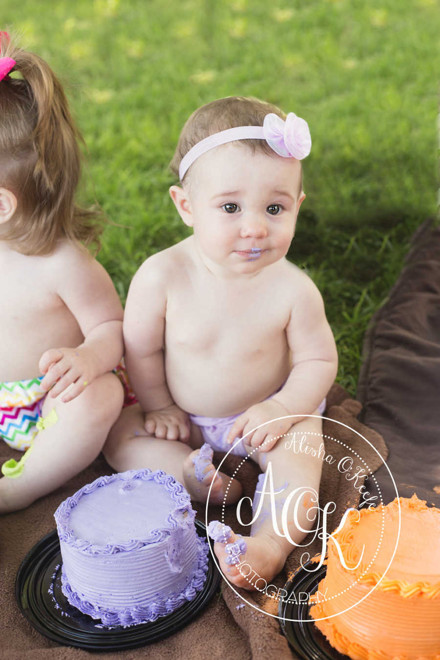 Infant with pink headband with bow sits with purple cake in front of her