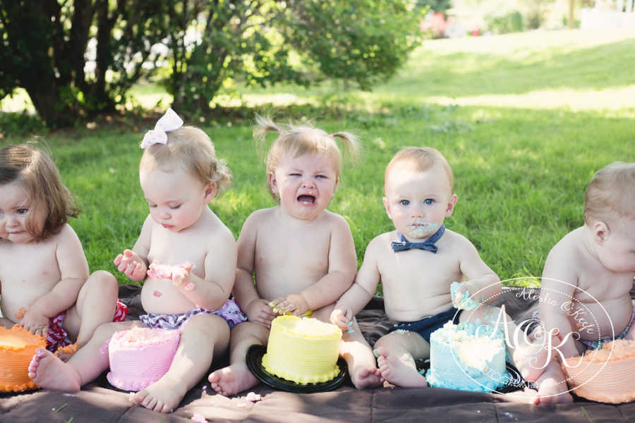 Infant crying with yellow cake in front of her besides other infants with cake