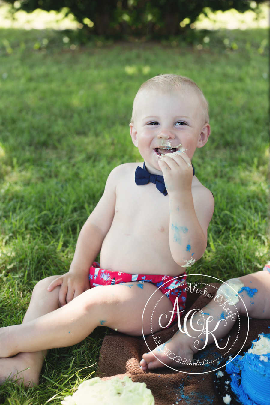 Shirtless infant with bowtie on smiles as he takes a bite of cake with his hand