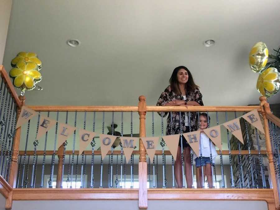 Woman stands with daughter at birth parents home that have sign on banister saying, "Welcome home"