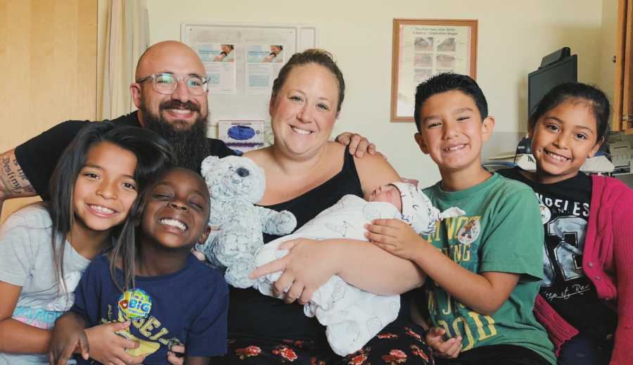 Foster mother holds her newborn in hospital beside four foster kids and husband