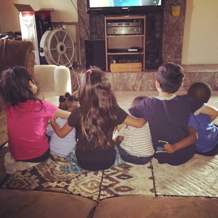 Six foster siblings sit on floor of home with arms around each other watching tv
