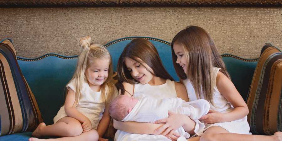 Three young girls sit on couch holding and looking at newborn who was abandoned by mother in hospital