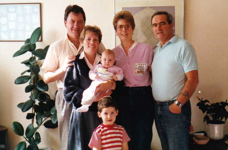 Husband and wife stand smiling with adopted baby daughter and toddler son beside couple