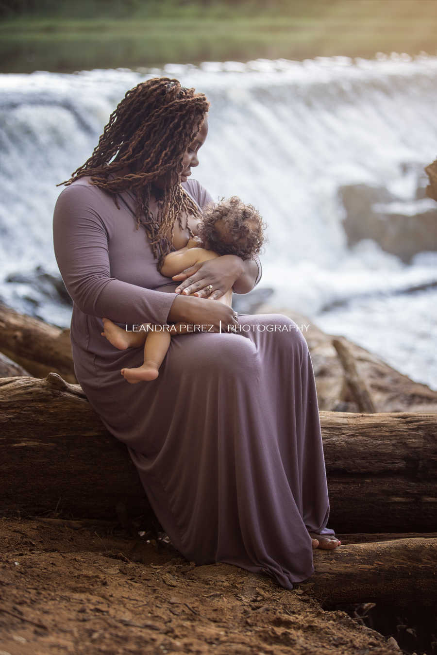 Mother sits on log beside river breastfeeding
