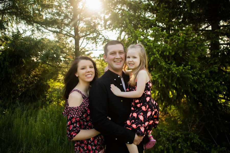 Young man smiles holding young daughter with fiancee standing behind him