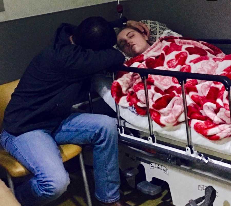 Teen with Gastroparesis lies in hospital bed asleep while boyfriend sits next to her rubbing her head