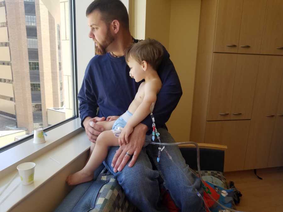 Little boy with cancer sitting on fathers lap as they watch what happens out the window