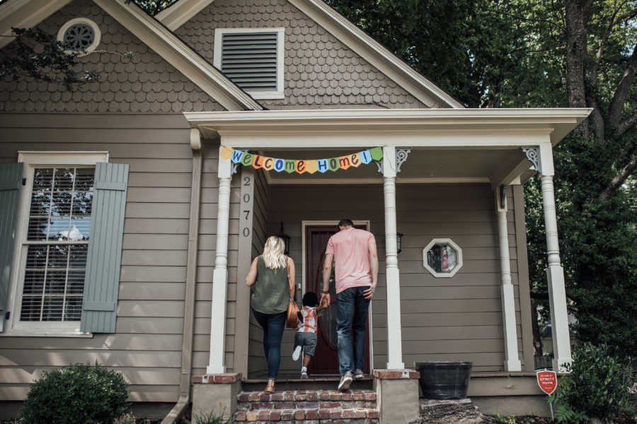 Adoptive parents walk into home with toddler son with sign saying ,"Welcome home" on front porch