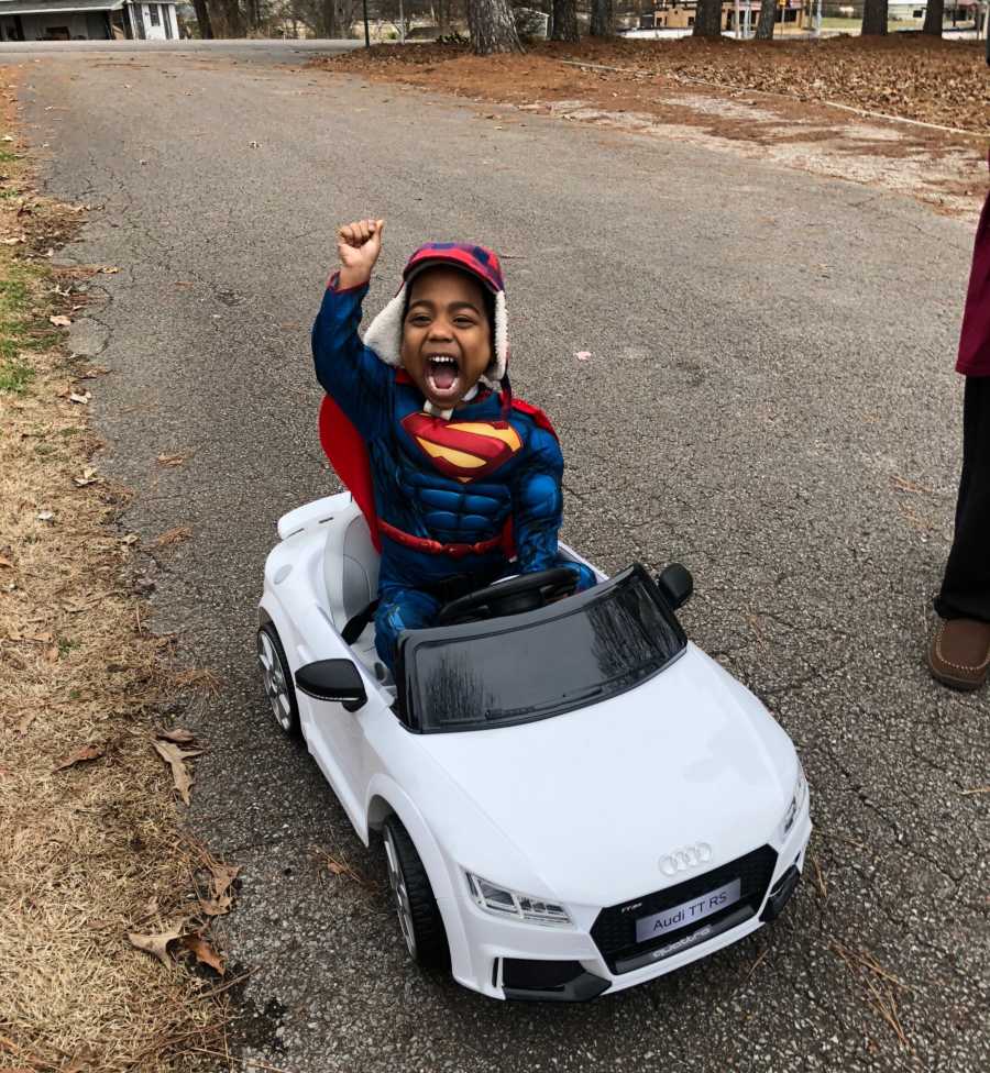 Adopted little boy in superman suit sits in toy electric car with one arm in air