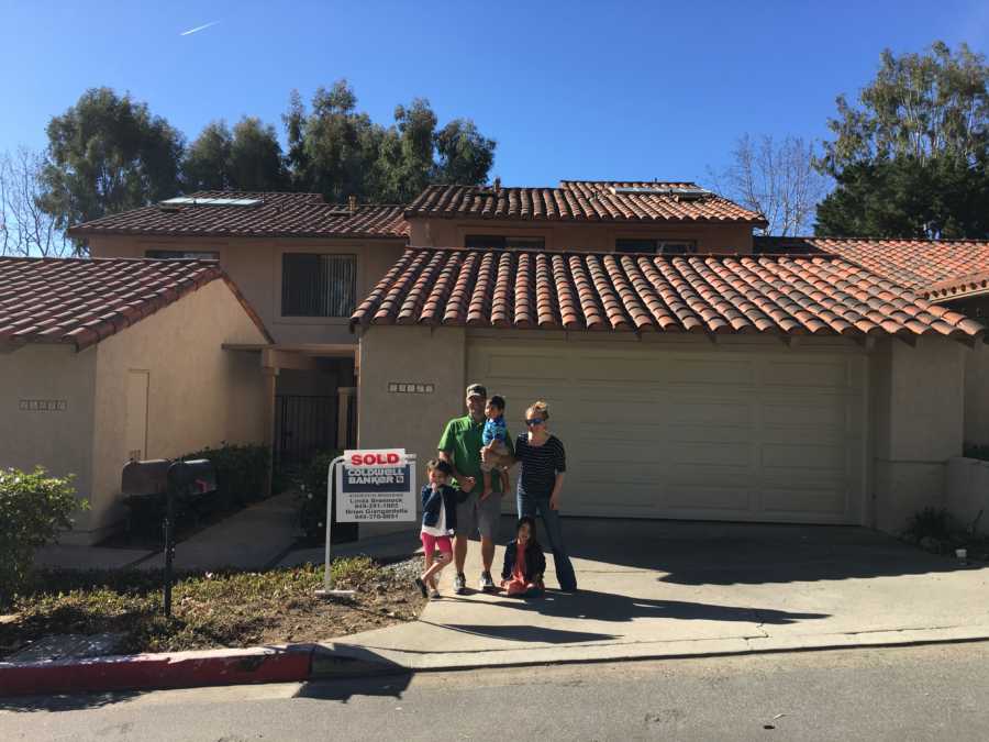 Husband and wife stand in front of new home next to sold sign with three foster kids