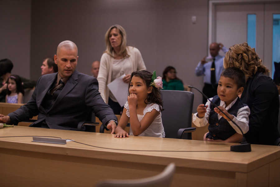 Father sits next to foster daughter and son at adoption court with people in background watching