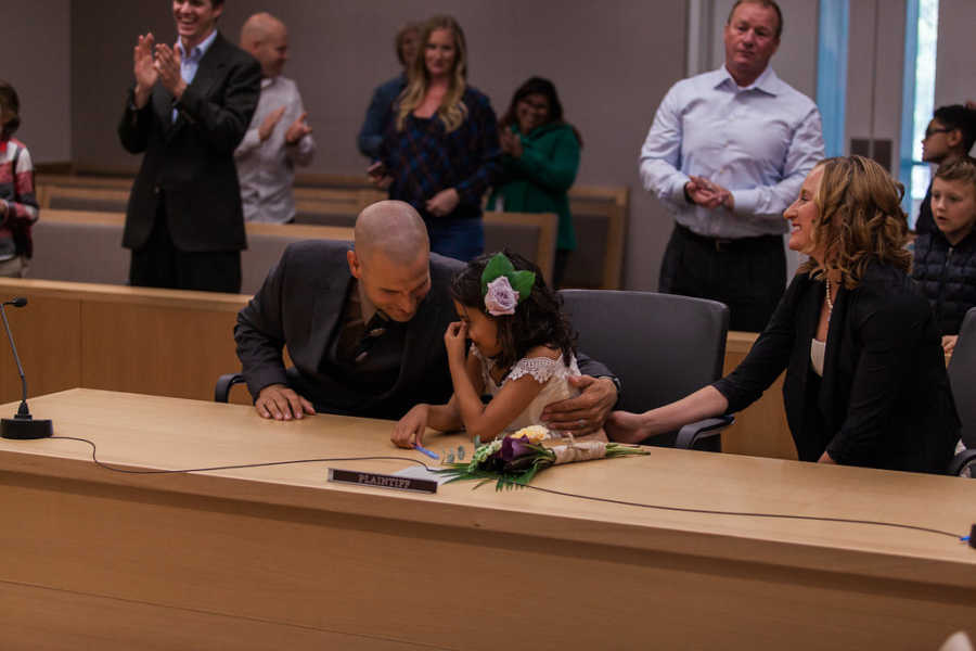 Husband and wife hold foster daughter who is now adopted at adoption court with people behind clapping