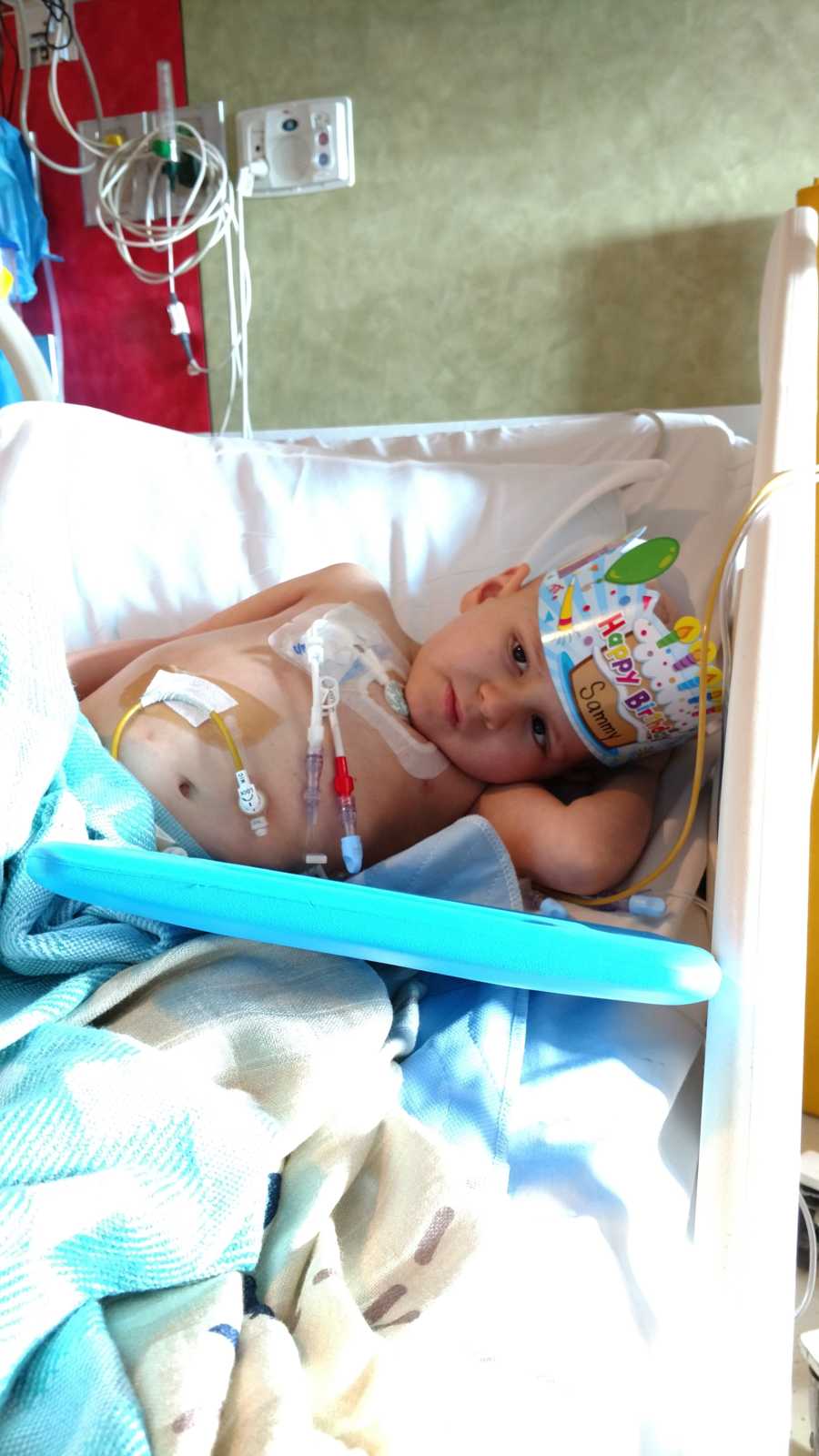 Little boy with brain cancer lying in hospital bed with Birthday crown on and wires attached to body