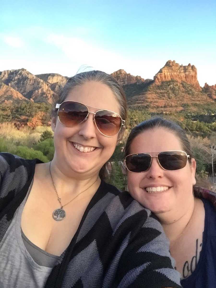 Same sex couple smile in selfie with mountains in the background