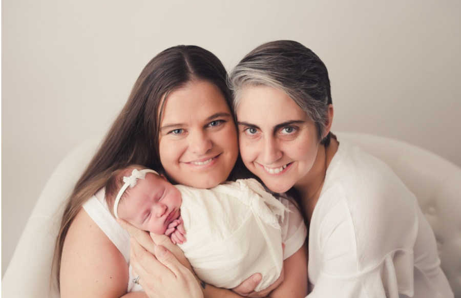 Mothers hold newborn baby swaddled in white blanket who was conceived through IVF