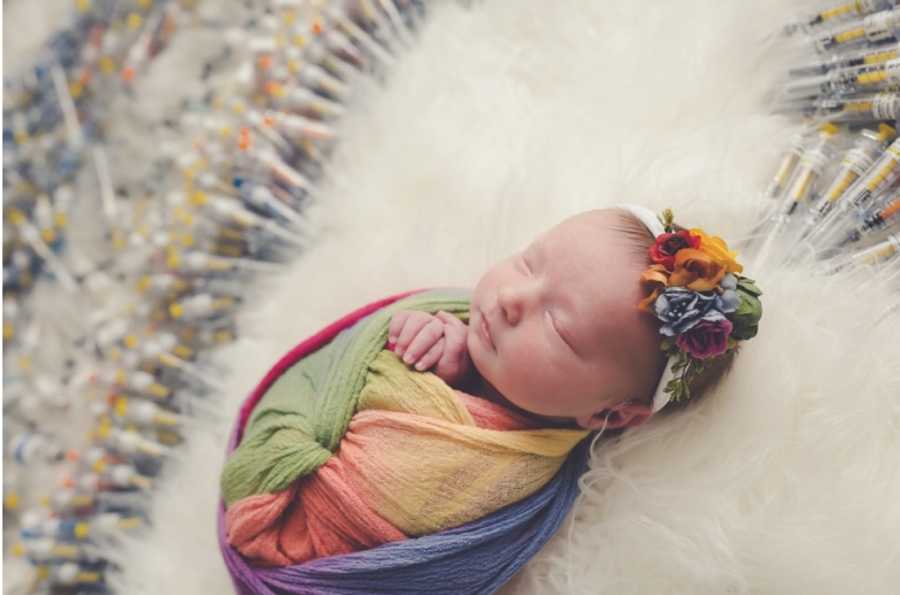 Newborn of two mothers lay swaddled in rainbow blanket surrounded by IVF needles