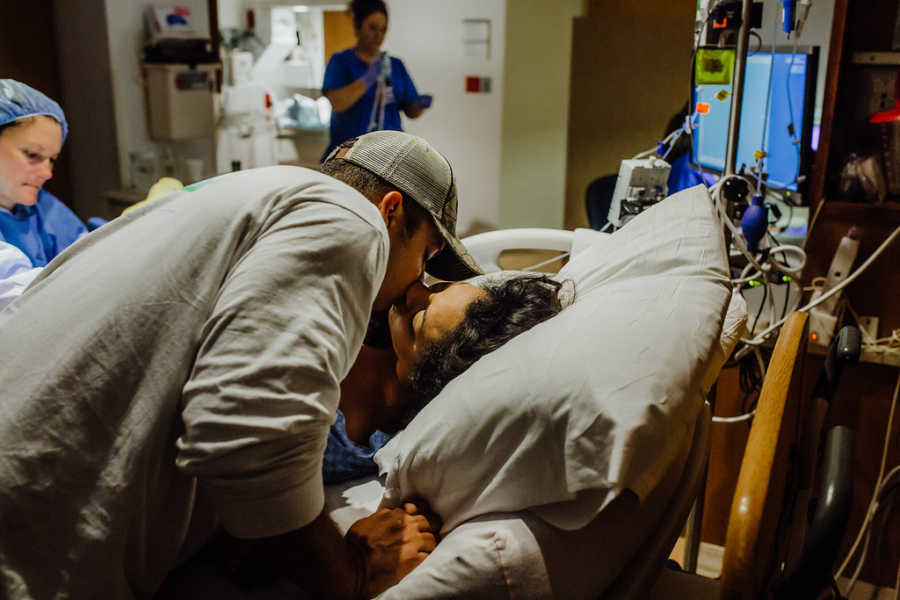 Husband leans over to kiss wife lying in hospital bed who struggled to get pregnant with third child
