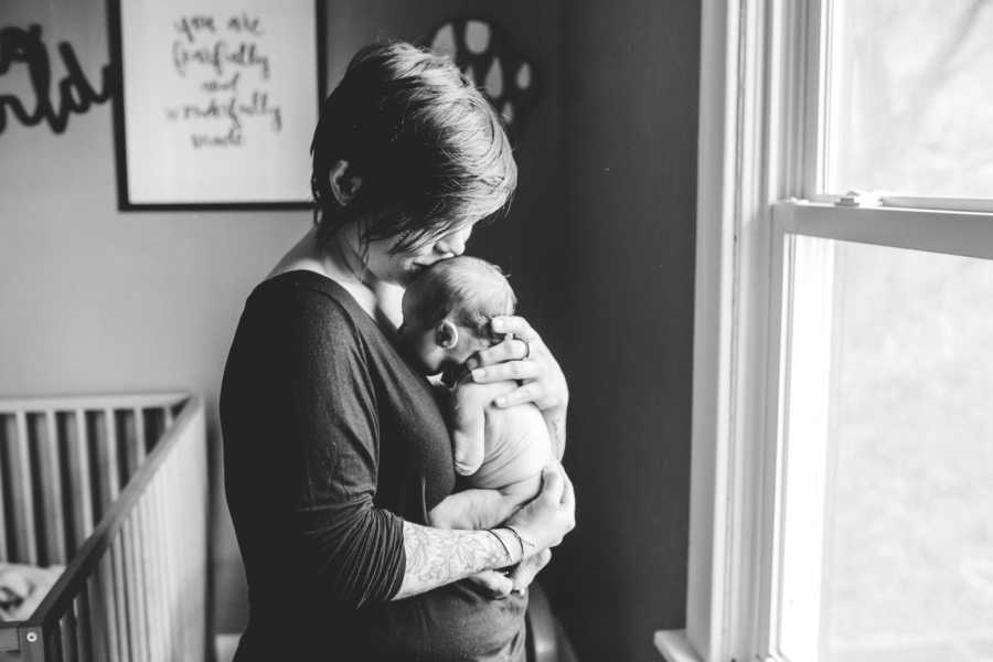 Foster mom stands holding newborn baby kissing it's forehead near window in nursery