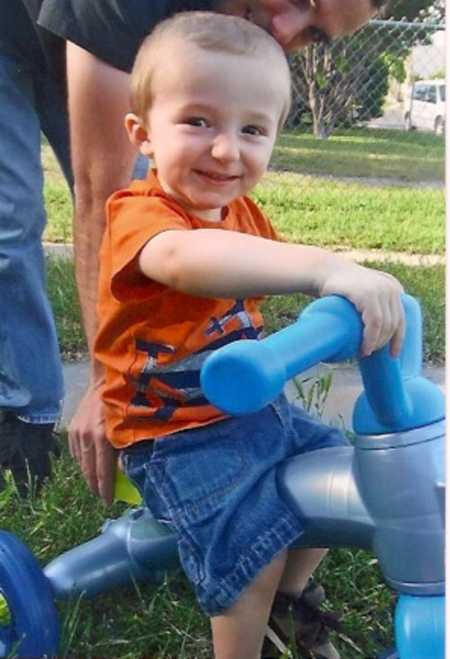 Little boy whose mother had him at seventeen smiles while sitting on plastic tricycle