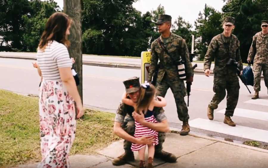 Soldier father bends town to hug his oldest daughter while his wife stands in front of him holding younger daughter