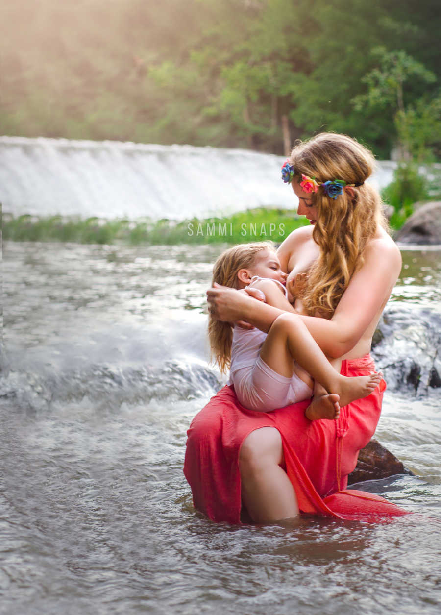 Mother sits on rock in river breastfeeding her three year old
