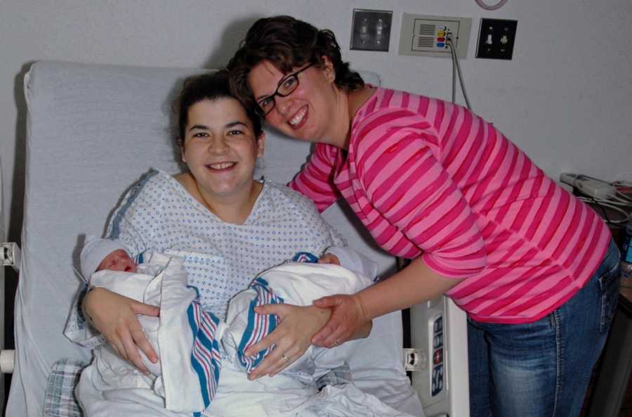 Mother leans over hospital bed where surrogate sits holding two newborns