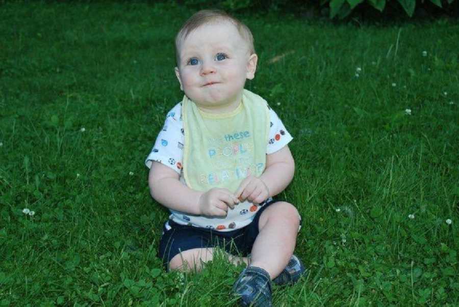 Infant boy wearing a bib smiles as he sits in grass