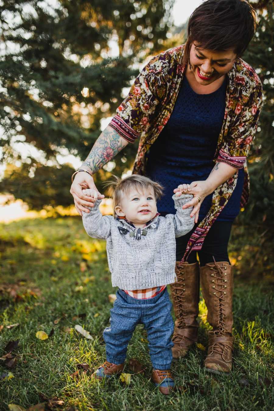 Woman stands holding adopted toddlers hands as he stands in front of her