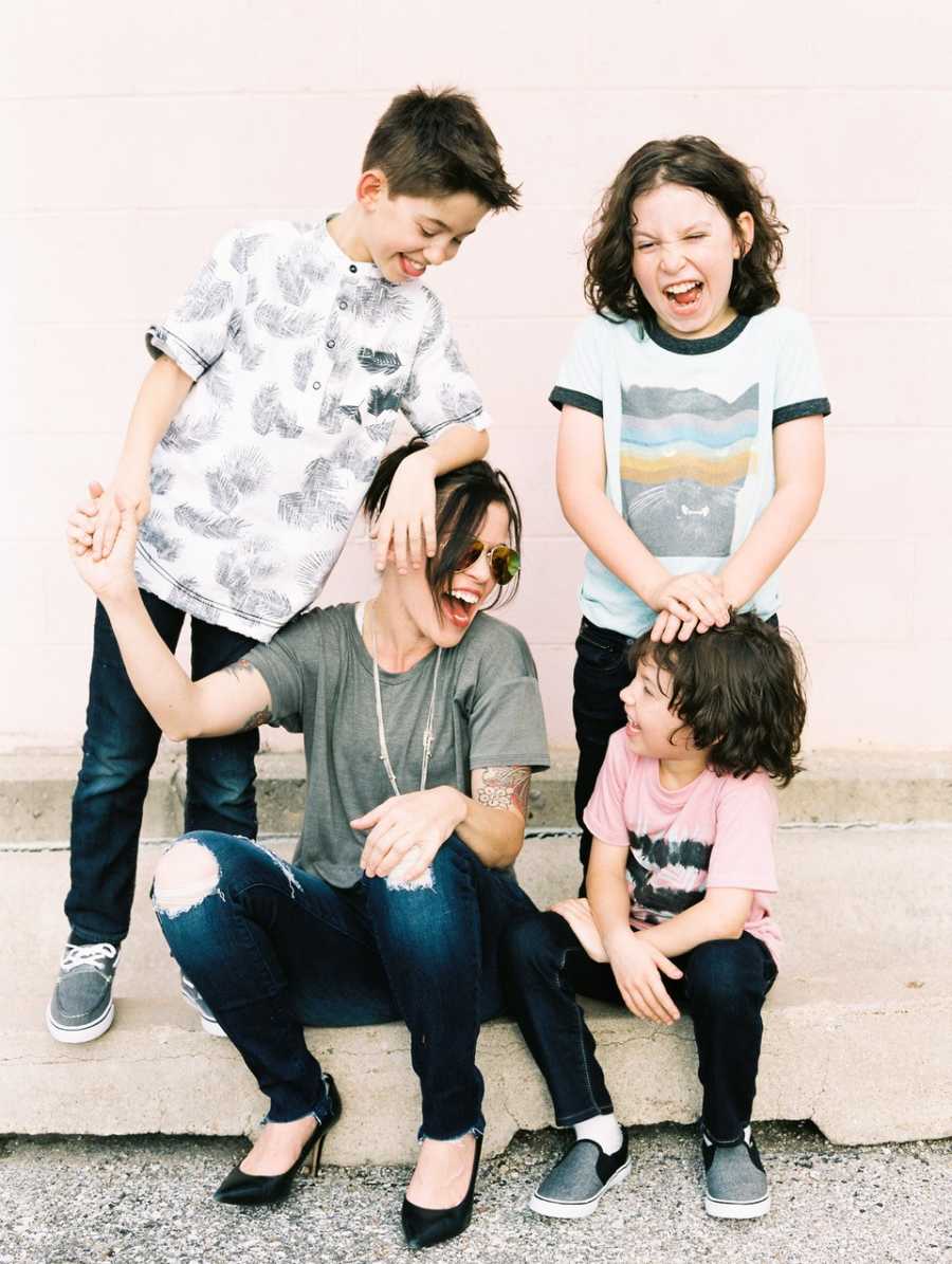 Mother sits on curb beside youngest son with two oldest, one of which has heart issues, stands over them