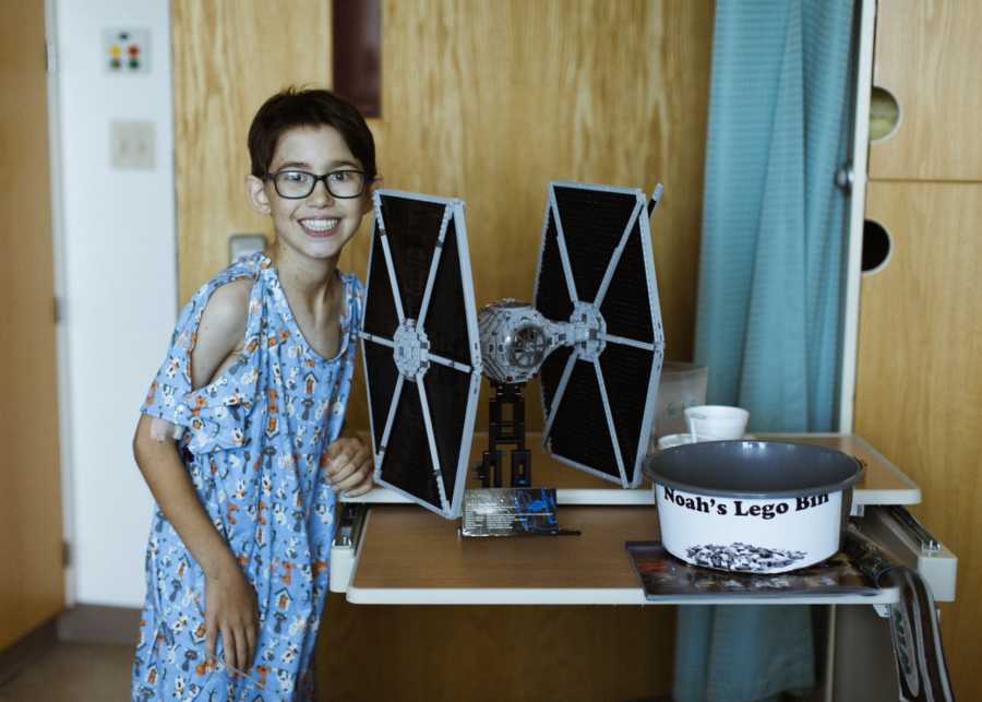 Young boy with heart condition stands smiling in hospital beside Star Wars structure made of legos