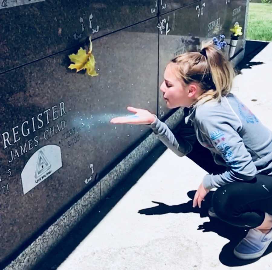Daughter crouches down on the ground to blow glitter on her father's gravestone