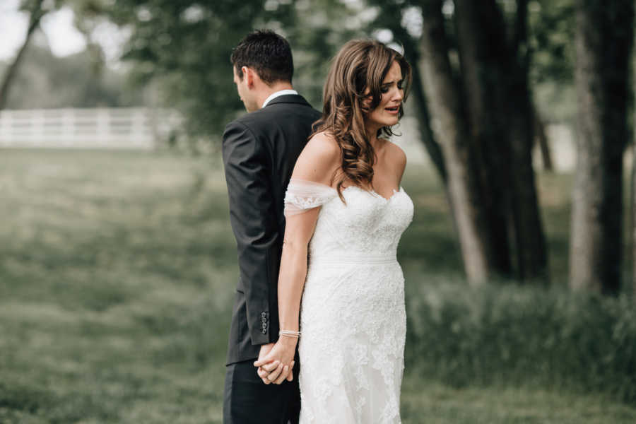 Bride and groom stand back to back holding hands as bride cries