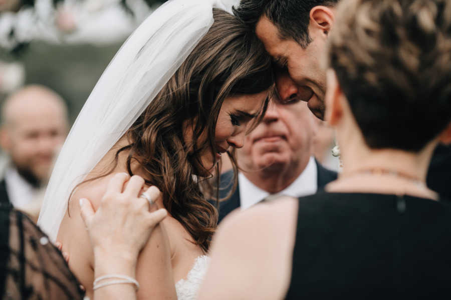 Bride and groom stand head to head sobbing at wedding with people watching