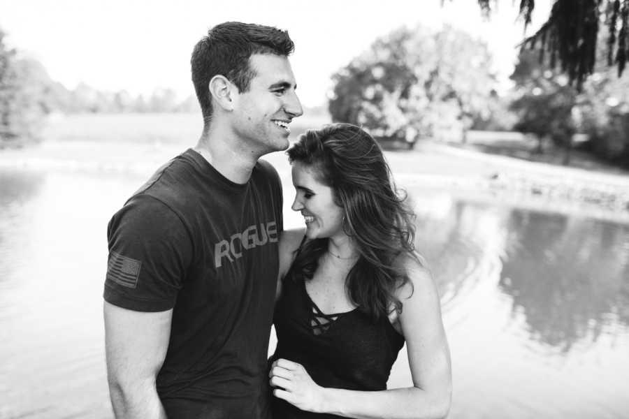 Couple stands arm in arm smiling in front of body of water