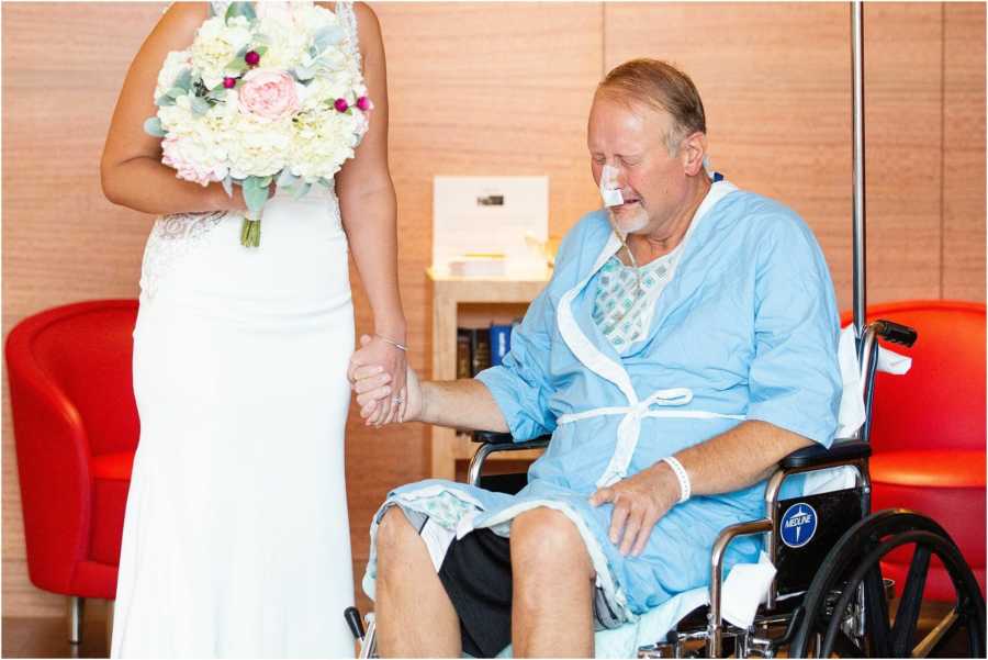 Father with pancreatitis sits crying in wheel chair holding daughter's hand who is a bride