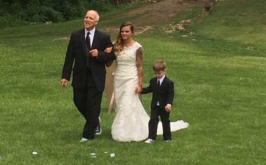 Bride with tattoos all over her arms walks with father and son through grass