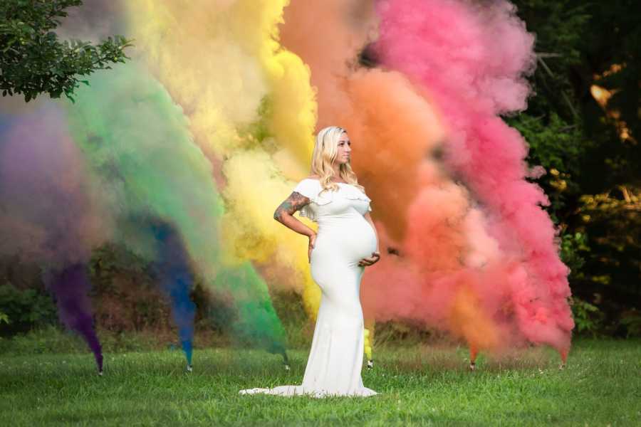 Pregnant woman who suffered child loss stands in white dress holding stomach with rainbow smoke behind her