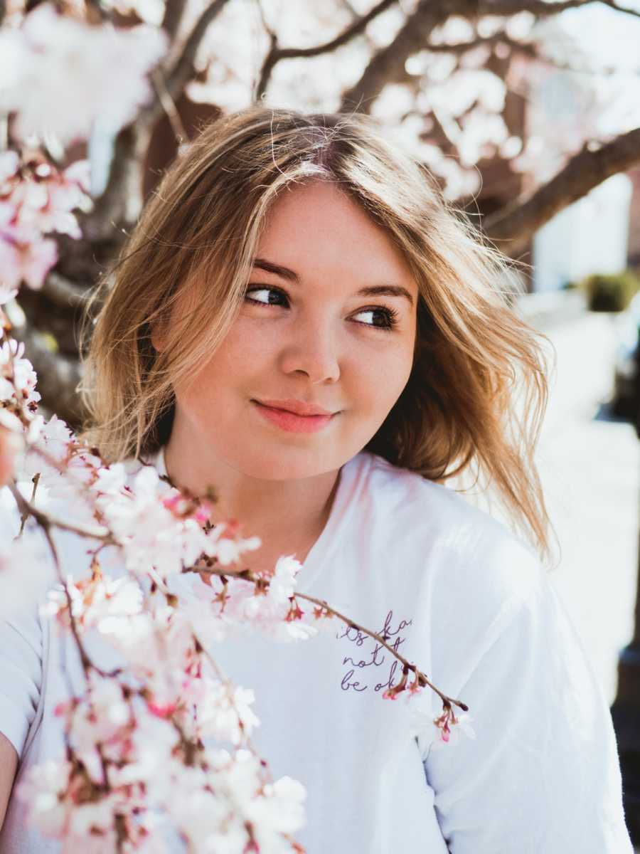 Woman recovering from suicide attempt smiles during photoshoot while sitting next to a tree with pink flowers