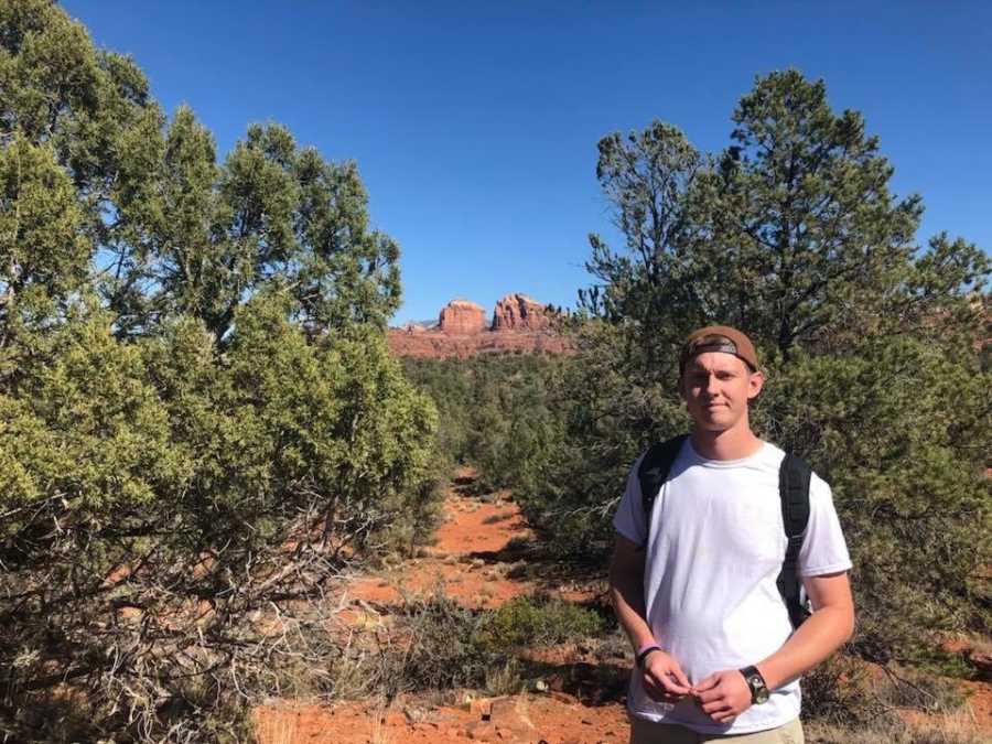 Young man in white t-shirt with brown hat and backpack takes a photo in front of huge rocks behind him