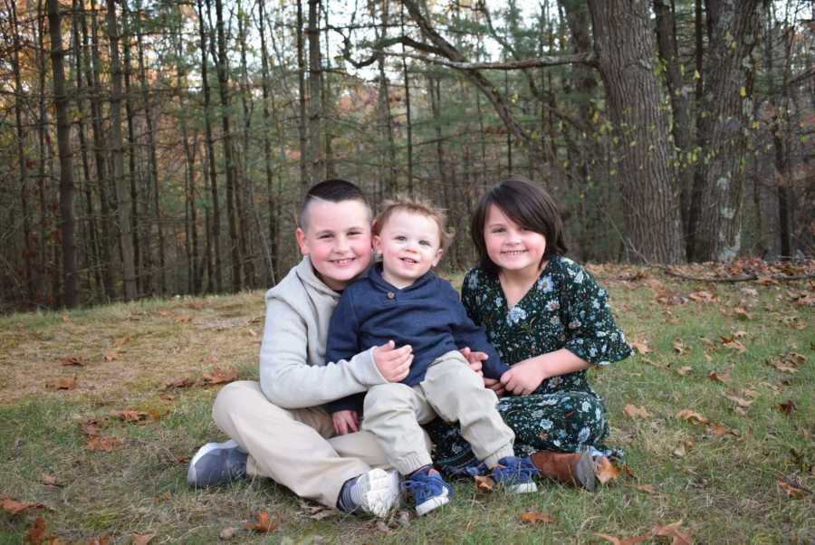 Little boy who is now a girl sits on ground outside with older and younger brother