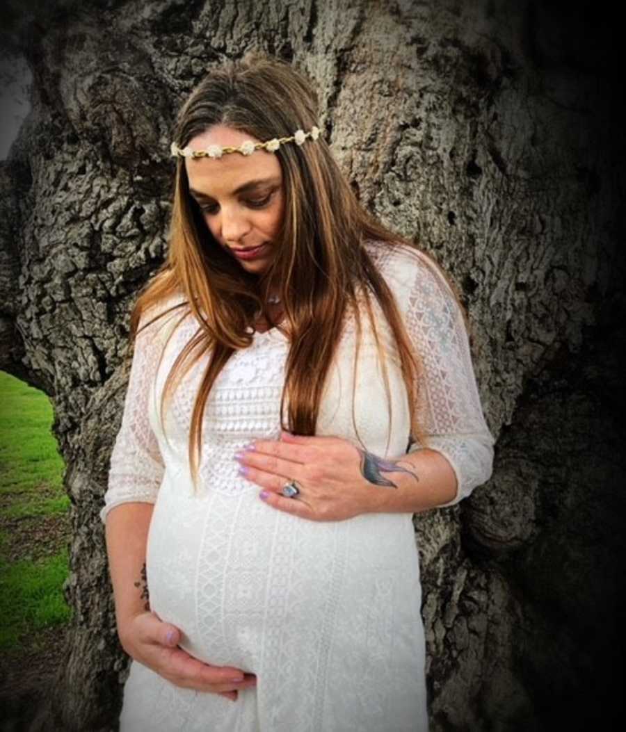 Pregnant woman who couldn't believe she was pregnant stands in white dress and flower crown holding her stomach
