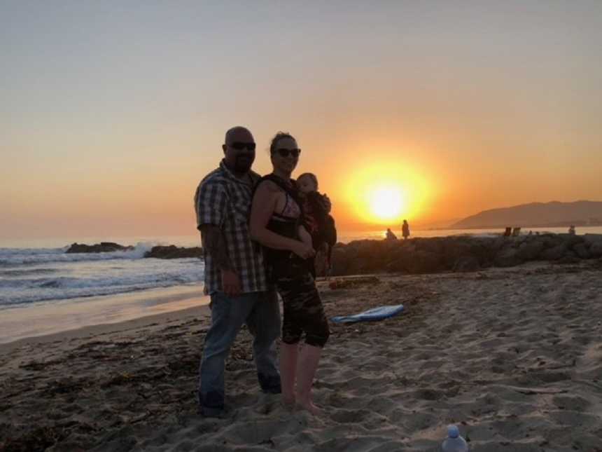 Husband stands behind wife who has baby swaddled to her on beach at sunset
