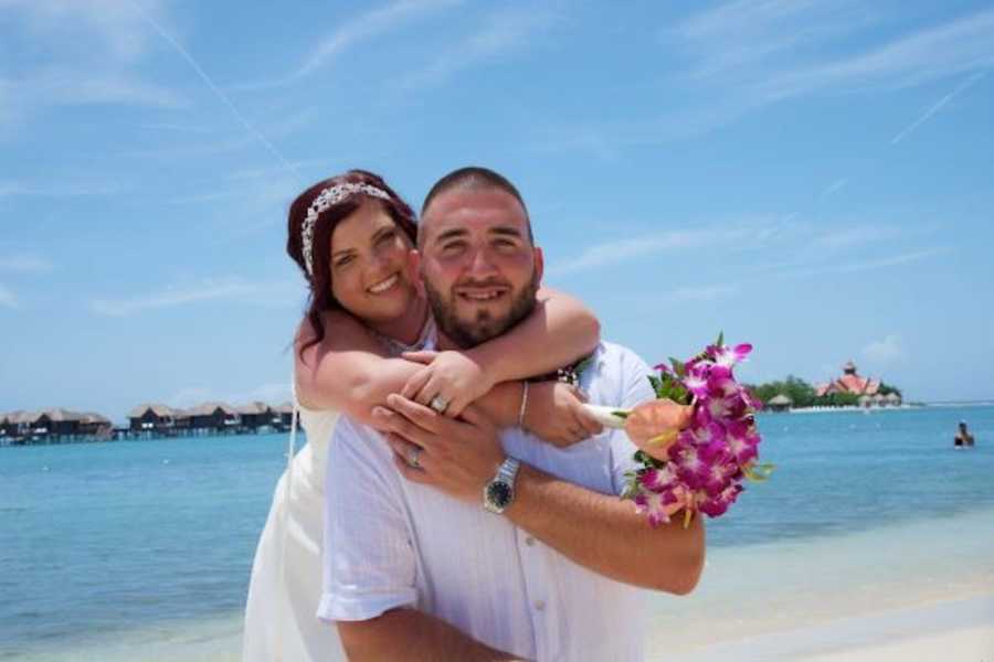 Bride smiles standing behind husband with her arms wrapped around him on beach