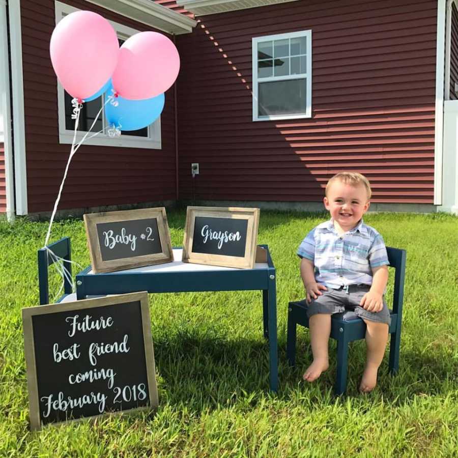 Young son sits at a children's play table outside next to a sign announcing his parents are having a second child