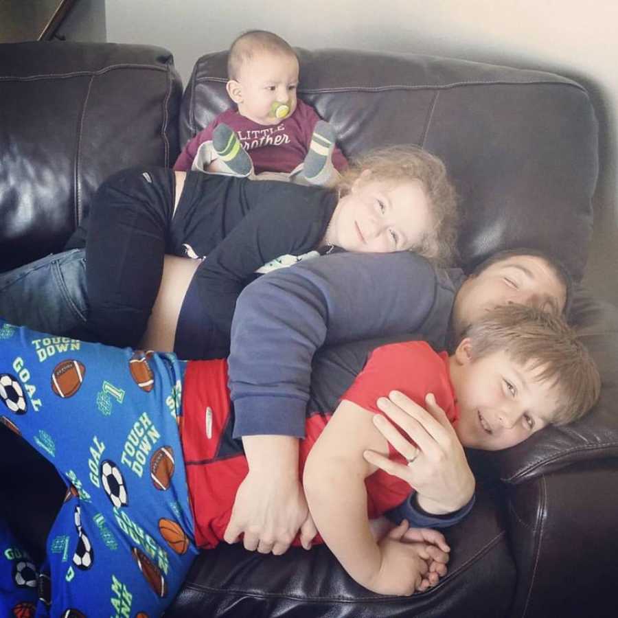 Father lays on couch with oldest son beside him, daughter on top, and baby on top of her