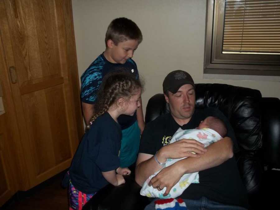 Father sits on black leather couch with new born in his arms with older children standing next to him