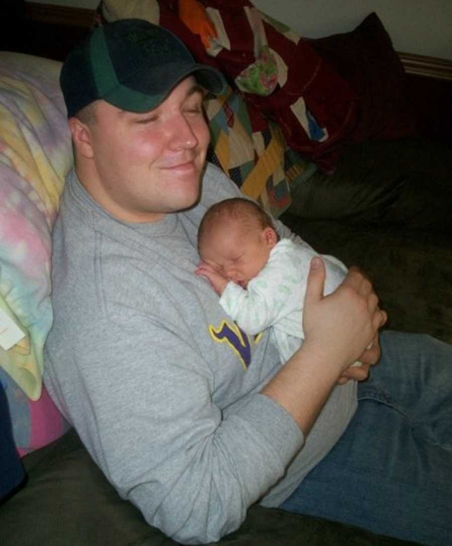 Father sits smiling on couch holding his newborn son on his chest