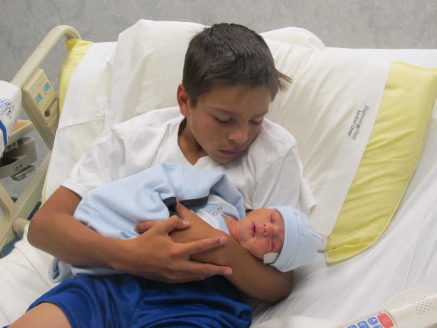 Young boy holds newborn baby in hospital bed that belongs to his father and stepmom